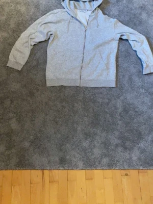 Ljusgrå zip hoodie med huva - En enkel och stilren ljusgrå hoodie med dragkedja framtill och huva med dragsko. Hoodien har långa ärmar och ribbade muddar vid ärmslut och nederkant. Perfekt för en avslappnad look och passar till det mesta.