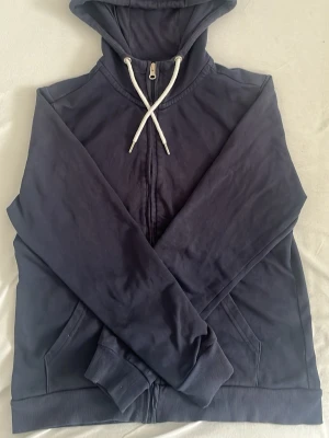 Mörkblå hoodie med dragkedja - Snygg mörkblå hoodie med dragkedja framtill och vita dragsnören i huvan. Hoodien har ribbade muddar vid ärmslut och nederkant samt två fickor fram. Perfekt för en avslappnad och stilren look.