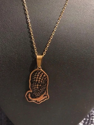 Palestina keffya Guldigt halsband med siluettberlock - Unikt halsband i guldfärgad metall med en siluettberlock som föreställer en person med flätat hår. Kedjan är tunn och stilren, och berlocken har ett detaljerat mönster på framsidan medan baksidan är slät. Perfekt accessoar för dig som gillar personliga smycken.