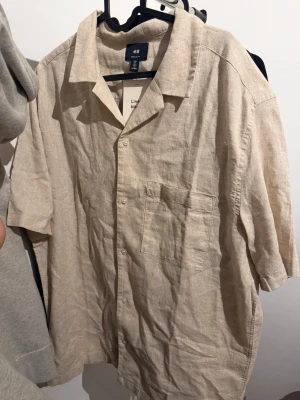 Beige linneskjorta från H&M - Säljer en beige kortärmad skjorta från H&M i linnemix. Skjortan har klassisk krage, bröstficka och knappar framtill. Perfekt för varma dagar och enkel att styla med jeans eller shorts.