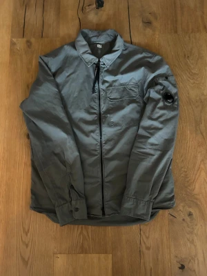 Mörkgrön cp company overshirt med linsdetalj - Stilren grå overshirt med dragkedja framtill och bröstficka. Unik linsdetalj på vänster ärm ger en cool touch. Skjortkrage och knappslut vid ärmslut. Tillverkad i ett tunt, mjukt material som passar perfekt för lager på lager.