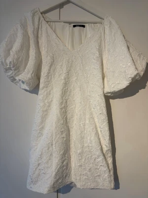 Vit klänning med puffärmar - Kort vit klänning med djup V-ringning och stora puffärmar. Klänningen har ett vackert strukturmönster i tyget som ger en exklusiv känsla. Materialet är tjockt och brokadliknande med diskret glans. Perfekt för dig som vill sticka ut med en trendig och elegant look. Använd endast en gång men är i helt nytt skick. Väldigt fin klänning som passar i många sammanhang.