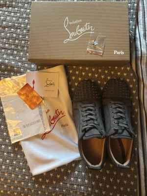 Gråa loubs sneakers med spikes med röd sula - Säljer ett par gråa sneakers från med klassiska röda sulor och nitar på tån. Skorna är i mocka och har snörning framtill. Medföljer originalkartong, dustbag. Perfekta för dig som vill sticka ut med exklusiv stil. OBS en spike/ knit saknas på höger sko som man kan se på sista bilden.