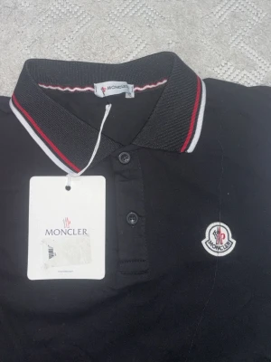 Svart pikétröja från Moncler - Svart pikétröja från Moncler med klassisk krage och knäppning framtill. Kragen har röda och vita ränder som snygg detalj. Moncler-logga broderad på bröstet. Tillverkad i 100% bomull för en skön känsla.
