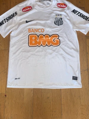 Vit Santos FC matchtröja Nike - Säljer en vit Santos FC matchtröja från Nike med Dri-FIT material. Tröjan har klubbmärke, flera sponsorloggor och texten 'Menino da Vila desde 1912' i nacken. Perfekt för fotbollsträning eller att bära på läktaren.