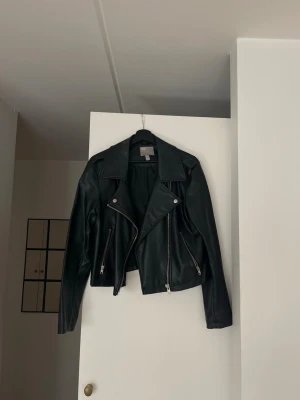 Svart klassisk skinnjacka - Passar mig som normalt bär storlek 38.Snygg svart skinnjacka med silvriga dragkedjor och breda slag. Jackan har två framfickor med dragkedja och detaljerade sömmar. Perfekt för dig som vill ha en tidlös bikerstil i garderoben.