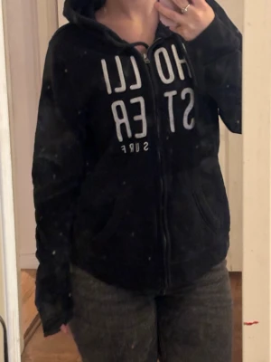 Svart hoodie med tryck och dragkedja - Säljer en svart hoodie med vit text på bröstet och dragkedja framtill. Hoodien har huva med snörning och två fickor fram. Perfekt för en avslappnad och cool stil. Materialet känns mjukt och bekvämt. Står strl L men liten i strl så passar väst xs/s se bilderna( smutsig spegel)