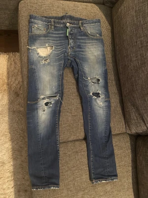 Blå slitna dsquared2 jeans - Säljer ett par blå dsquared2 jeans med kraftiga slitningar och hål på både fram- och baksida. Jeansen har en tvättad look, fem fickor och råa benslut. Perfekta för dig som gillar en edgy och trendig stil.