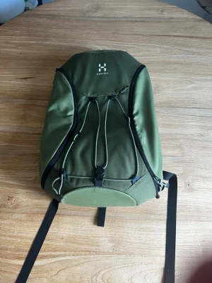Olivgrön ryggsäck från Haglöfs - Snygg olivgrön ryggsäck från Haglöfs i slitstarkt Cordura-material. Ryggsäcken har svarta detaljer, dragkedjor och justerbara remmar. Praktisk design med snörning framtill och flera fack för enkel organisering. Perfekt för skola eller äventyr.