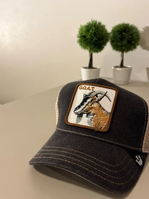 GOAT Keps - Snygg svart truckerkeps med vit mesh baktill och ett broderat patch framtill med en get och texten 'GOAT'. Kepsen har böjd skärm och kontrastsömmar. Perfekt för dig som vill sticka ut med en unik accessoar.