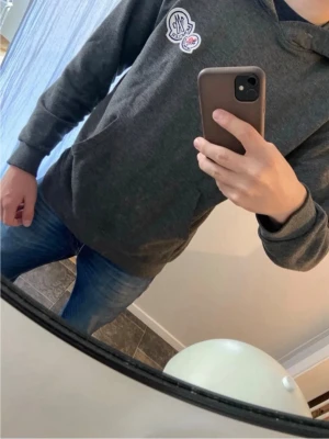 Moncler hoodie  - Snygg mörkgrå hoodie med huva och två små tryck på bröstet. Hoodien har klassisk känguruficka framtill och ribbade muddar vid ärmslut och nederkant. Perfekt för en avslappnad och trendig look. M MEN PASSAR OCKSÅ S 