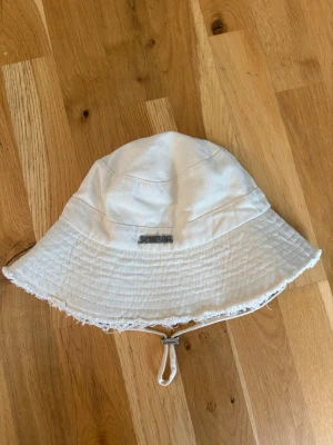 Vit bucket hat med texten Jacquemus - Vit bucket hat med texten Jacquemus med broderad logga framtill. Hatten har råa kanter och justerbart hakband. Tillverkad i bomull och har en avslappnad, klassisk form som passar perfekt till sommaren.