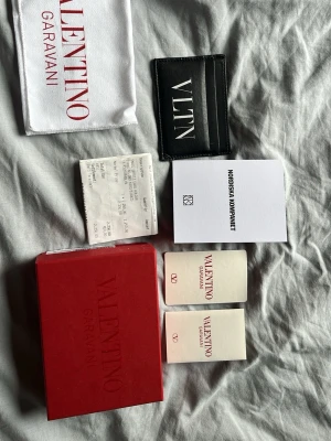 Svart Valentino korthållare i läder - Snygg svart korthållare från Valentino Garavani i äkta läder med vit VLTN-logga på framsidan. Kommer med originalförpackning, dustbag och äkthetskort. Perfekt för dig som vill ha en stilren och exklusiv accessoar till dina kort.