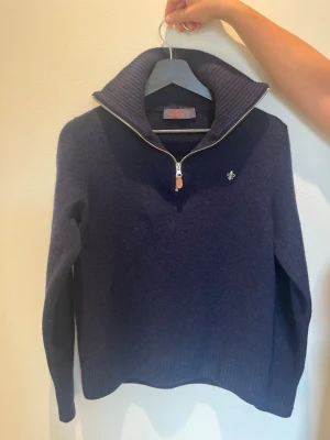 Mörkblå stickad tröja med halvzip från Morris - Mysig mörkblå stickad tröja med halvzip och hög krage. Tröjan har ett litet broderat emblem på bröstet och ribbade muddar vid ärmslut och nederkant. Perfekt för kyliga dagar och enkel att styla till olika outfits. Storleken är M men passar även S.