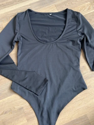 Grå långärmad body med djup urringning - En stilren grå body med långa ärmar och djup rund urringning. Tillverkad i ett mjukt och stretchigt material som sitter tajt mot kroppen. Perfekt att matcha med jeans eller kjol för en clean look.