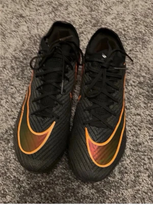 Svarta fotbollsskor med orange detaljer - Snygga fotbollsskor i svart med stora orangea detaljer och Nike-logga på ovansidan. Skorna har snörning och en slitstark sula med Air Zoom-teknologi för extra komfort och grepp på planen. Perfekta för dig som vill sticka ut på fotbollsplanen.