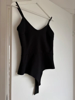 Svart body med smala axelband - Snygg svart body med smala axelband och v-ringad hals. Modellen har en enkel och stilren design med markerad midja och mjukt, stretchigt tyg. Perfekt att matcha med jeans eller kjol för en trendig look. Finns en liten defekt som inte syns när plagget är på (se bild 3)