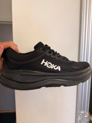 Svarta Hoka sneakers med tjock sula - Snygga svarta sneakers från Hoka med kraftig, vadderad sula och ventilerande mesh-ovandel. Skorna har klassisk snörning och Hoka-logga i vitt på sidan. Perfekta för dig som gillar streetwear och komfort i vardagen.