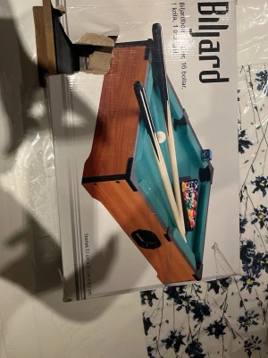 Mini biljardbord  - Säljer ett mini biljardbord i trä med grön filt, två köer, 16 färgglada bollar, triangel, krita och en svart låda. Perfekt för spel hemma eller på festen. Bordet är kompakt och lätt att ställa undan när det inte används.