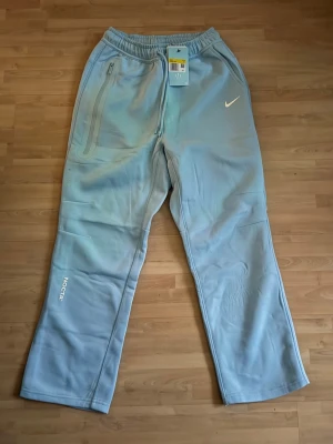 Nike Nocta Byxor - Ljusblå Nike NOCTA mjukisbyxor från  med raka ben, resår i midjan och dragkedjeficka på ena sidan. Swoosh-logga framtill och litet NOCTA-tryck på benet. Sköna och stilrena, perfekta för en chill look. Kom gärna med frågor och prisförslag!