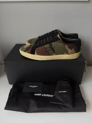 Yves saint laurent skor - Saint Laurent Court Classic Camo Sneakers – Strl 40  Säljer ett par stilrena Saint Laurent Court Classic i camouflage. Väldigt snygg modell som är enkel att matcha och ger en exklusiv look.  Skick: Mycket fint skick, inga tecken på användning Färg: Camo/Grön, svart och beige. Storlek: 40 Nypris: ca 7200kr Ingår: Originalkartong, dustbag och tillbehör enligt bild.