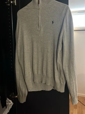 Grå halvzip tröja från Ralph Lauren - Snygg grå långärmad tröja från Ralph Lauren med halv dragkedja och hög krage. Tröjan har den klassiska broderade loggan på bröstet och ribbade muddar vid ärmslut och nederkant. Perfekt för en clean och stilren look.