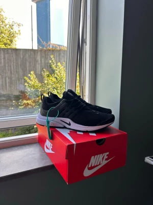 Svarta Nike sneakers med vit sula - Snygga svarta Nike sneakers med vit sula och mesh-överdel för bra andningsförmåga. Skorna har en sportig design med klassisk Nike-logga på sidan och elastiska snören för enkel påtagning. Perfekta för dig som gillar streetwear och sportig stil.