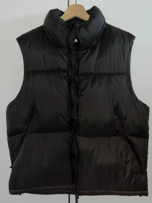 Svart dunväst från Zara - Snygg svart dunväst från Zara med hög krage och dragkedja framtill. Västen har två sidofickor och en puffig, quiltad design som ger en modern look. Perfekt att slänga över en hoodie eller tröja för extra värme under kyliga dagar.