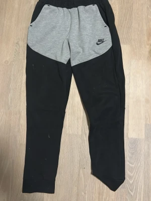 Nike svart-grå nike tech mjukisar - Nike mjukisbyxor i svart med grå panel framtill och stora fickor. Byxorna har resår i midjan, smal passform och en dragkedjeficka bak. Perfekta för chill eller träning, med klassisk Nike-logga på ena fickan.