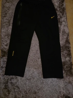 Svarta NOCTA x Nike mjukisbyxor - Svarta mjukisbyxor från Nike och NOCTA med gul logga och detaljer. Byxorna har raka ben, dragsko i midjan och en dragkedjeförsedd ficka. Perfekta för en chill och sportig look.