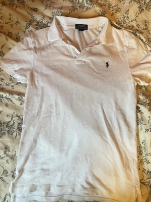 Vit pikétröja från Polo Ralph Lauren - Klassisk vit pikétröja från Polo Ralph Lauren med liten broderad logga på bröstet. Tröjan har krage, korta ärmar och knappar vid halsen. Perfekt till både jeans och shorts för en clean och stilren look.