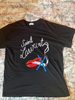Saint Laurent Paris SS18 ”NO SMOKING” t-shirt - Svart t-shirt från Saint Laurent med coolt tryck framtill som visar röda läppar, blått kryss och vit text. Klassisk rund hals och korta ärmar. Perfekt för dig som vill sticka ut med en unik design. Tillverkad i mjuk bomull för skön känsla.