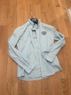 Ljusblå skjorta från Tommy Hilfiger - Säljer en ljusblå skjorta från Tommy Hilfiger med klassisk button-down krage och broderad logga på bröstet. Skjortan har långa ärmar och vita knappar framtill samt vid ärmsluten. Perfekt till jeans eller chinos.