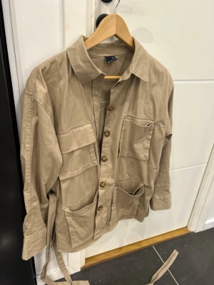 Beige overshirt med fickor och bälte - Stilren beige overshirt med fyra stora fickor framtill, knäppning med bruna knappar och tillhörande bälte i midjan. Skjortkrage och långa ärmar med knapp vid ärmslut. Tillverkad i ett mjukt bomullsliknande material, perfekt för lager på lager.