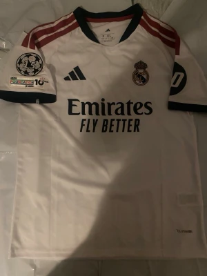 Real Madrid matchtröja Vini Jr. 7 - Vit Real Madrid matchtröja från Adidas med Vini Jr. och nummer 7 på ryggen. Tröjan har röda och mörkblå detaljer, klubbmärke och sponsortryck framtill samt Champions League-märke på ärmen. Perfekt för fotbollsfans!