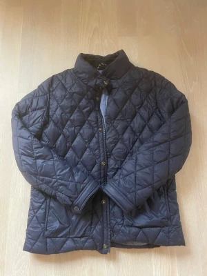 Mörkblå quiltad vindjacka - Snygg mörkblå quiltad vindjacka med tryckknappar och ståkrage. Jackan har ett rutmönster och är lätt vadderad, perfekt för blåsiga dagar. Klassisk design som funkar till det mesta och ger en stilren look.
