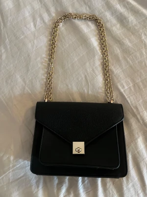 Svart axelväska med guldkedja - Stilren svart axelväska med guldfärgad kedjerem. Väskan har ett elegant vridlås framtill och är gjord i ett slätt, läderliknande material. Perfekt för dig som gillar en klassisk och trendig look. Justerbart band