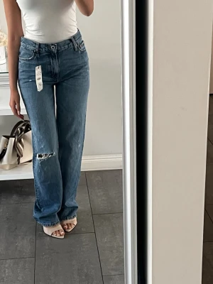 Blå raka jeans med slitning - Säljer ett par blå raka jeans med hög midja och en snygg slitning på ena knät. Jeansen har klassisk femficksmodell och är tillverkade i denim. Perfekta för en avslappnad och trendig look.