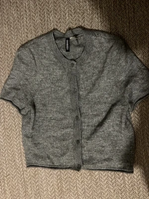 Grå kortärmad kofta från Boohoo - Kortärmad grå kofta från Boohoo med rund halsringning och knappar framtill. Ribbstickade detaljer vid hals, knappslå och ärmslut. Perfekt att styla över ett linne eller t-shirt för en chill look.