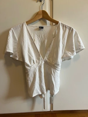 Vit omlottopp med knytband - Snygg vit omlottopp med korta ärmar och knytband i midjan. Toppen har en djup v-ringning och är gjord i mjuk bomull, perfekt för en fräsch och enkel look. Passar till både jeans och kjol för en trendig stil. Välj gärna instabox