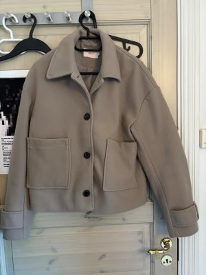 Beige kappa - Snygg beige kappa från Nelly med stora framfickor och klassisk krage. Jackan har knäppning med svarta knappar och uppvikta ärmslut. Perfekt för dig som gillar en stilren och enkel look. lite nopprig men inget som syns tydligt