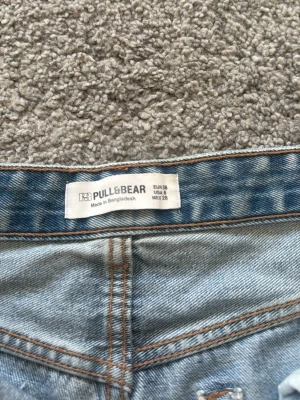 Ljusa jeansshorts från Pull&Bear - Säljer ett par ljusblå jeansshorts från Pull&Bear och slitna detaljer. Shortsen har råa kanter, flera slitningar framtill och klassiska fem fickor. Perfekta för sommaren och ger en avslappnad look.