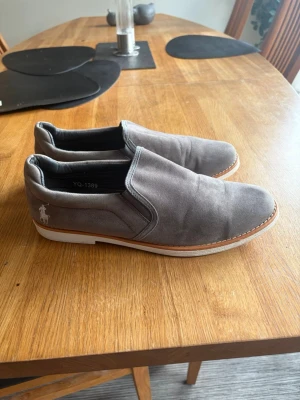 Gråa loafers i mocka - Ralph Lauren - Säljer ett par stilrena gråa loafers i mocka med vit sula och bruna detaljer längs sulkanten. Skorna har en enkel slip-on design och en diskret logga på hälen. Perfekta för dig som gillar en clean och avslappnad look.
