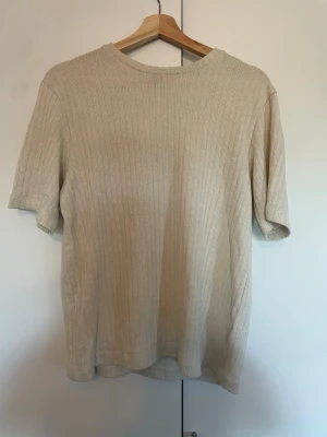 Beige ribbad t-shirt - En stilren beige t-shirt med ribbad struktur och rund hals. T-shirten har korta ärmar och är tillverkad i ett mjukt, stickat material som ger en avslappnad men ändå trendig look. Perfekt för dig som gillar minimalistisk stil.