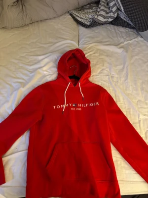 Röd hoodie från Tommy Hilfiger - Snygg röd hoodie från Tommy Hilfiger med vit logga och flagga på bröstet. Tillverkad i ekologisk bomull och har en klassisk känguruficka samt vita dragsnören i huvan. Använd fåtal gånger.