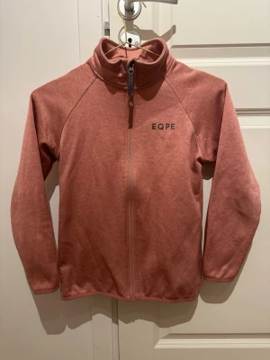 Rosa fleecejacka från EQPE - Mysig rosa fleecejacka från EQPE med hel dragkedja och hög krage. Perfekt för lager-på-lager och har en enkel, stilren design med EQPE-logga på bröstet. Tillverkad i mjukt fleecematerial som håller dig varm under kyliga dagar.