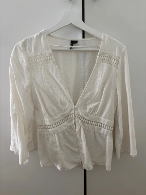 Vit blus med spetsdetaljer - Säljer en vit blus i boho-stil med djup v-ringning och dekorativa spetsdetaljer. Blusen har knappar framtill och vida, långa ärmar. Tillverkad i ett lätt, luftigt tyg som ger en avslappnad känsla.