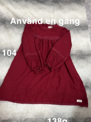 Vinröd långärmad klänning med spets - Vinröd klänning med långa ärmar och fina spetsdetaljer vid ärmslut, bröst och nederkant. Klänningen har en lös passform och rund halsringning. Perfekt för dig som gillar romantiska detaljer.🌸 Använd en gång. Storlek 104