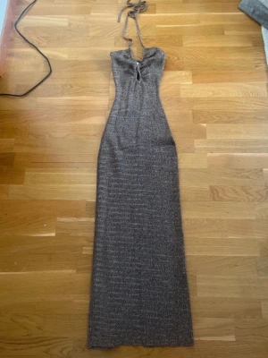 Adoore klänning  - Säljer en lång klänning i mörkbrun ton med smickrande passform. Klänningen har halterneck med snörning i nacken och en liten öppning framtill. Perfekt för dig som vill sticka ut med en elegant look. Sitter väldigt fint på