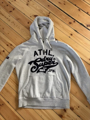 Grå hoodie med svart tryck - Snygg grå hoodie med stor svart Superdry-logga och japanska tecken på bröstet. Tröjan har huva med vita dragsnören och en klassisk magficka. Tillverkad i mjukt sweatshirtmaterial och har ribbade muddar vid ärmar och nederkant. Perfekt för en avslappnad streetwear-look.